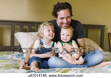 Stock Photograph - Father with sons (12-17 months, 4-5) sitting on bed. Fotosearch