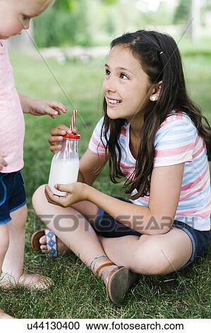 Stock Image - Girl (10-11) sharing bottle of milk with sister (2-3) in park. Fotosearch