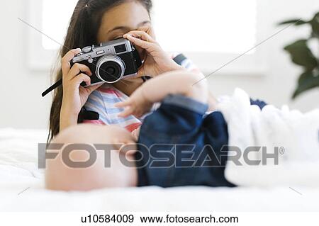 Stock Photo - Girl (10-11) taking photo of her small brother (12-17 months) lying on bed. Fotosearch