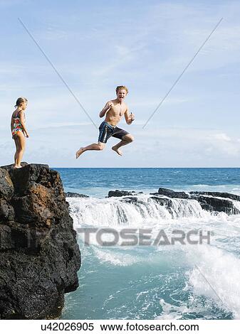 Stock Photography - Girl (6-7) standing on cliff and man jumping into sea. Fotosearch