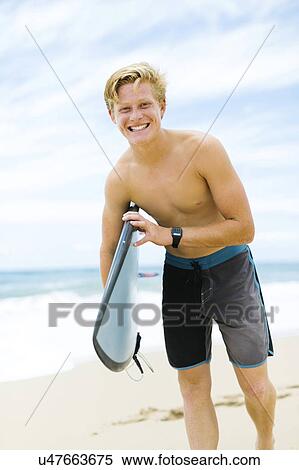 Stock Photography - Man standing on beach and holding surfboard. Fotosearch