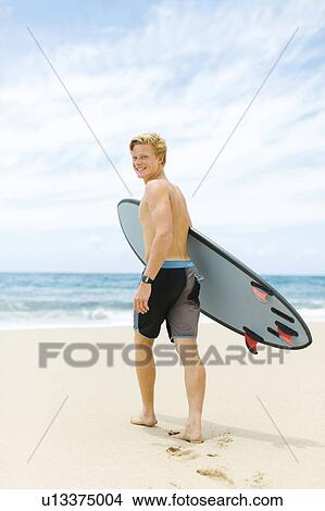 Picture - Man walking on beach and holding surfboard. Fotosearch