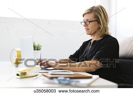 Stock Image - Mature woman in office typing on laptop. Fotosearch