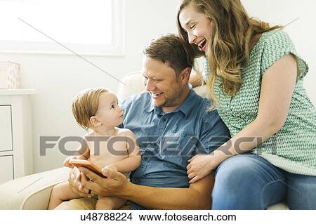 Picture - Mother and father with baby girl (12-17 months) sitting in living room and holding smart phone. Fotosearch