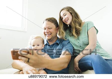 Mother and father with baby girl (12-17 months) sitting in living room and taking selfie View Large Photo Image Stock Image - Mother and father with baby girl (12-17 months) sitting in living room and taking selfie. Fotosearch
