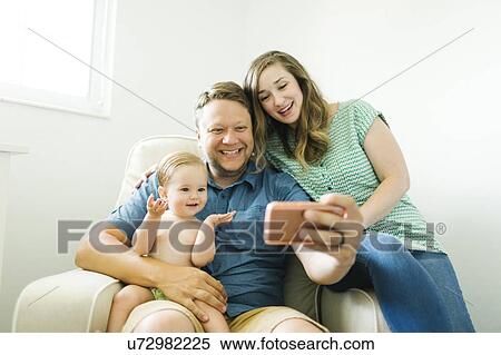Stock Photography - Mother and father with baby girl (12-17 months) sitting in living room and taking selfie. Fotosearch
