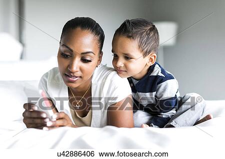 Stock Photograph - Mother and son (2-3) lying on bed and using smart phone. Fotosearch
