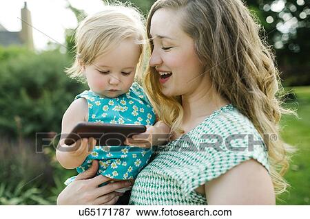 Stock Photo - Mother carrying daughter (12-17 months), daughter playing on smartphone. Fotosearch