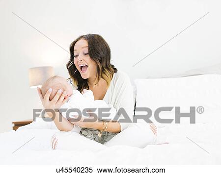 Mother sitting on bed carrying daughter (0-1 months) and laughing View Large Photo Image Stock Photography - Mother sitting on bed carrying daughter (0-1 months) and laughing. Fotosearch