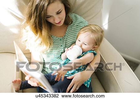 Stock Image - Mother with baby girl (12-17 months) sitting in living room and reading book. Fotosearch
