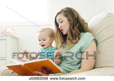 Stock Photo - Mother with baby girl (12-17 months) sitting in living room and reading book. Fotosearch