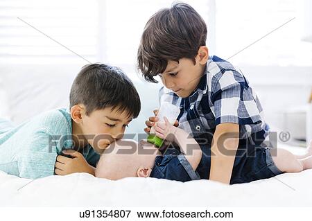 Stock Photo - Older brothers( 6-7, 8-9) feeding small brother (12-17 months) lying on bed. Fotosearch
