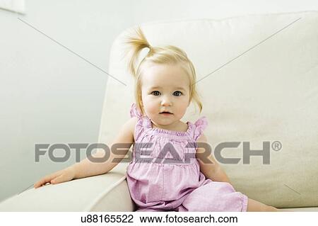 Stock Image - Portrait of baby girl (12-17 months) sitting on sofa. Fotosearch