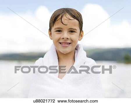 Stock Image - Portrait of boy (6-7) wrapped in towel by lake. Fotosearch