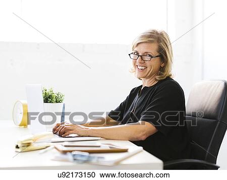 Stock Image - Portrait of Mature woman in office typing on laptop. Fotosearch
