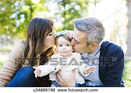 Stock Photo - Portrait of parents kissing daughter (12-17 months) in park. Fotosearch