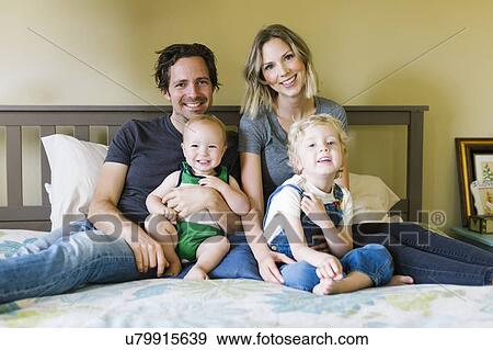 Stock Photo - Portrait of parents sitting with sons (12-17 months, 4-5) on bed. Fotosearch