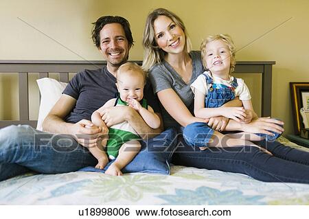 Stock Photograph - Portrait of parents sitting with sons (12-17 months, 4-5) on bed. Fotosearch