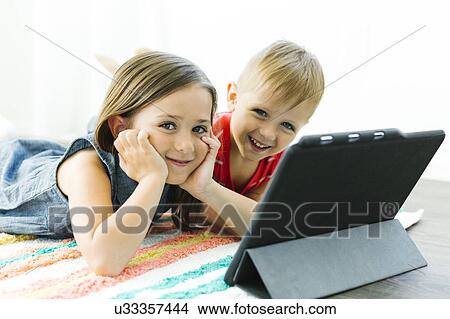 Picture - Portrait of siblings (2-3, 6-7) looking at camera and lying down on carpet next to digital tablet. Fotosearch