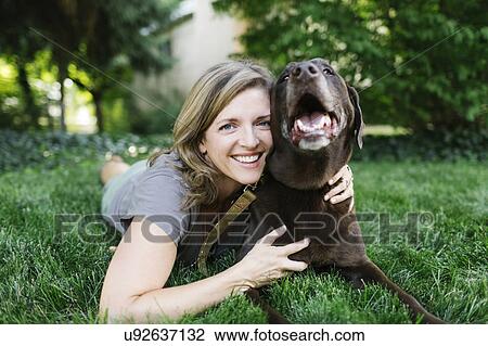 Stock Image - Portrait of smiling woman lying on grass with Labrador Retriever. Fotosearch