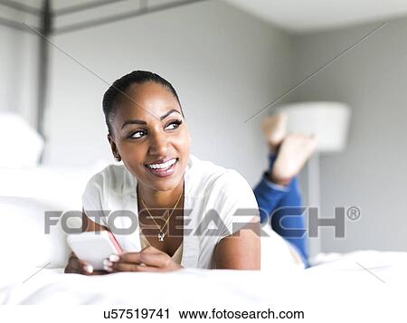 Stock Image - Portrait of woman lying on bed and holding smart phone. Fotosearch
