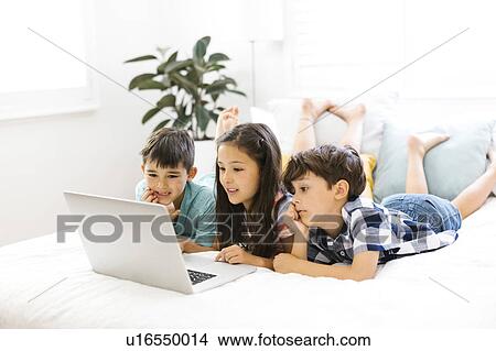 Picture - Siblings (10-11, 6-7, 8-9) lying in bed and using computer. Fotosearch