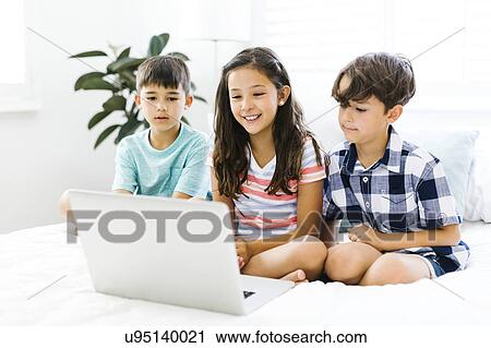 Stock Image - Siblings (10-11, 6-7, 8-9) sitting in bed and using computer. Fotosearch