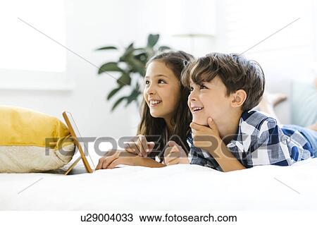 Stock Image - Siblings (10-11, 6-7) lying in bed and using tablet. Fotosearch