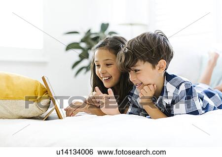 Stock Photograph - Siblings (10-11, 6-7) lying in bed and using tablet. Fotosearch