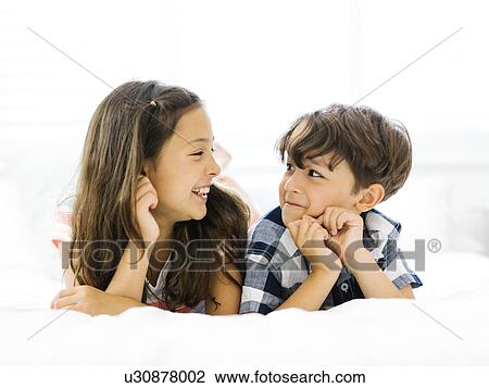 Stock Image - Siblings (10-11, 6-7) lying in bed side by side. Fotosearch