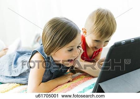 Stock Image - Siblings (2-3, 6-7) lying down on carpet and watching digital tablet. Fotosearch