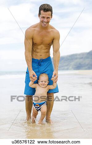 Stock Image - Son (12-17 months) learning to walk with father on beach. Fotosearch