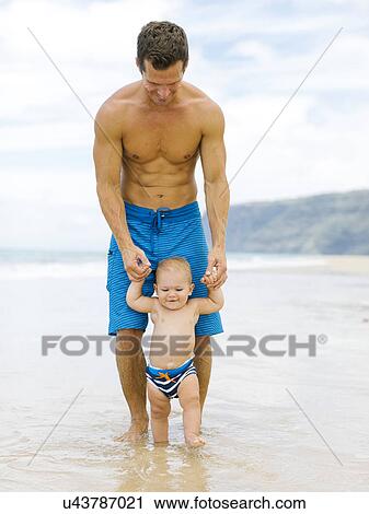 Stock Image - Son (12-17 months) learning to walk with father on beach. Fotosearch