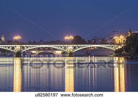 Stock Photo - Spain, Seville, Triana, Guadalquivir River and Puente de Isabel II at dusk. Fotosearch