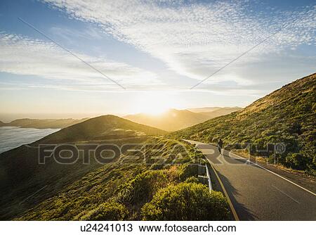 Stock Image - USA, California, San Francisco, California, Man walking on coastline road at sunset. Fotosearch