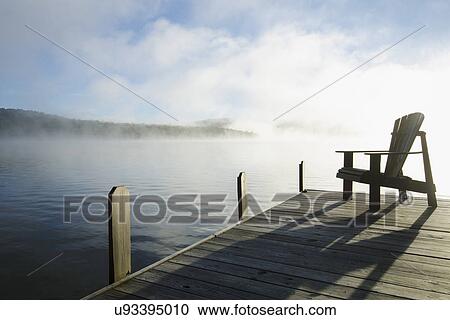 Stock Image - USA, New York, St. Armand, Lake Placid, Outdoor chair on pier by lake. Fotosearch