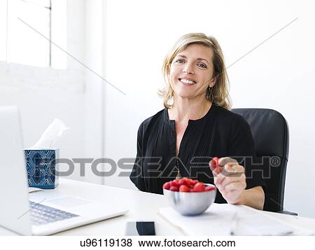 Stock Photo - Woman sitting in office, eating raspberries. Fotosearch