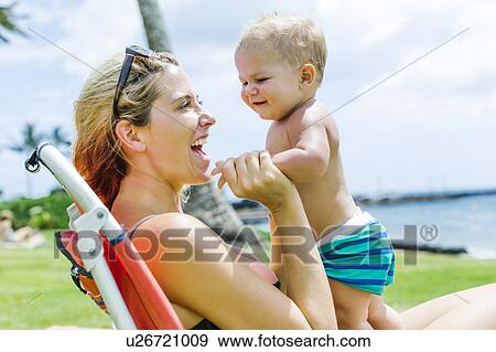 Stock Photo - Woman sitting under palm tree with little son (18-23 months). Fotosearch