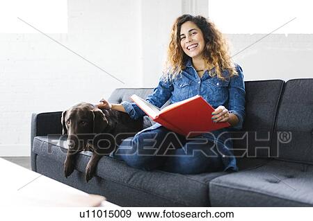 Stock Photo - Woman sitting with dog on sofa in living room and reading book. Fotosearch
