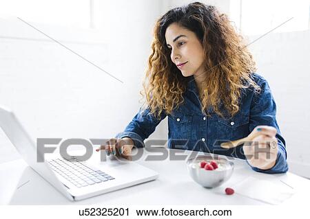 Stock Image - Woman using laptop and eating yoghurt. Fotosearch