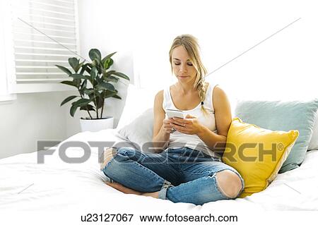 Young woman with mobile phone sitting on bed View Large Photo Image Stock Photo - Young woman with mobile phone sitting on bed. Fotosearch
