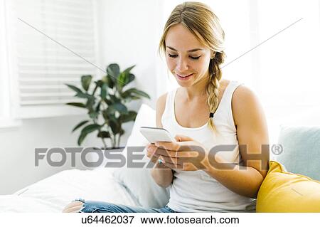 Stock Photo - Young woman with mobile phone sitting on bed. Fotosearch