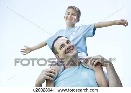 Stock Image - Father carrying son on shoulders, boy with his arms out.. Fotosearch