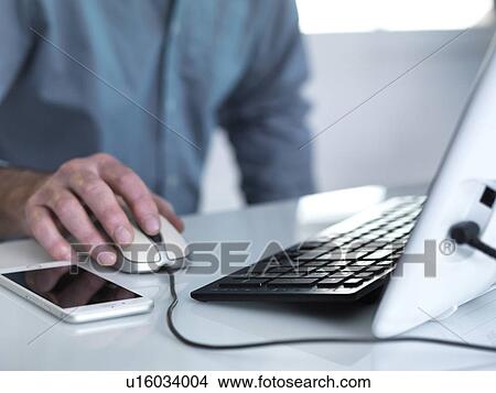 Picture - MODEL RELEASED. Computer use. Man working at PC with a mobile phone on the desk.. Fotosearch