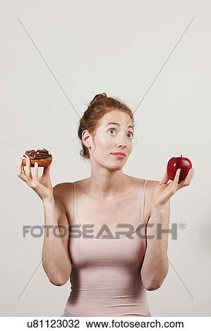 Stock Image - MODEL RELEASED. Indecisive woman holding apple and doughnut.. Fotosearch