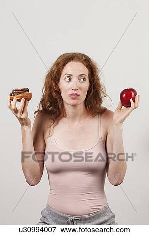 Stock Photo - MODEL RELEASED. Indecisive woman holding apple and doughnut.. Fotosearch