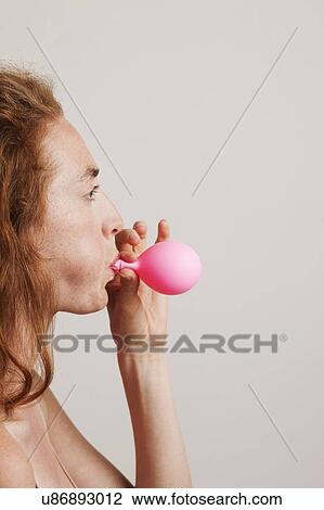 Stock Image - MODEL RELEASED. Profile of young woman with red hair blowing up pink balloon.. Fotosearch