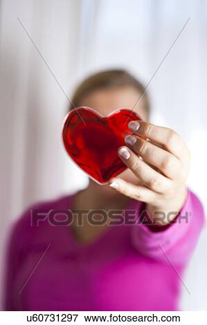 Stock Photo - MODEL RELEASED. Woman holding red heart in front of her face.. Fotosearch