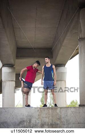 Stock Image - Two male athletes stretching under concrete bridge.. Fotosearch