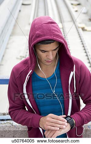 Young man wearing ear phones and hooded top, checking sports watch. View Large Photo Image Stock Photography - Young man wearing ear phones and hooded top, checking sports watch.. Fotosearch